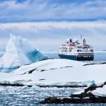 Un barco de expedición navega junto a un iceberg y unas rocas cubiertas de nieve bajo un cielo azul nublado