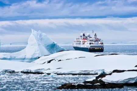 Ein Expeditionsschiff segelt unter einem bewölkten blauen Himmel an einem Eisberg und schneebedeckten Felsen vorbei