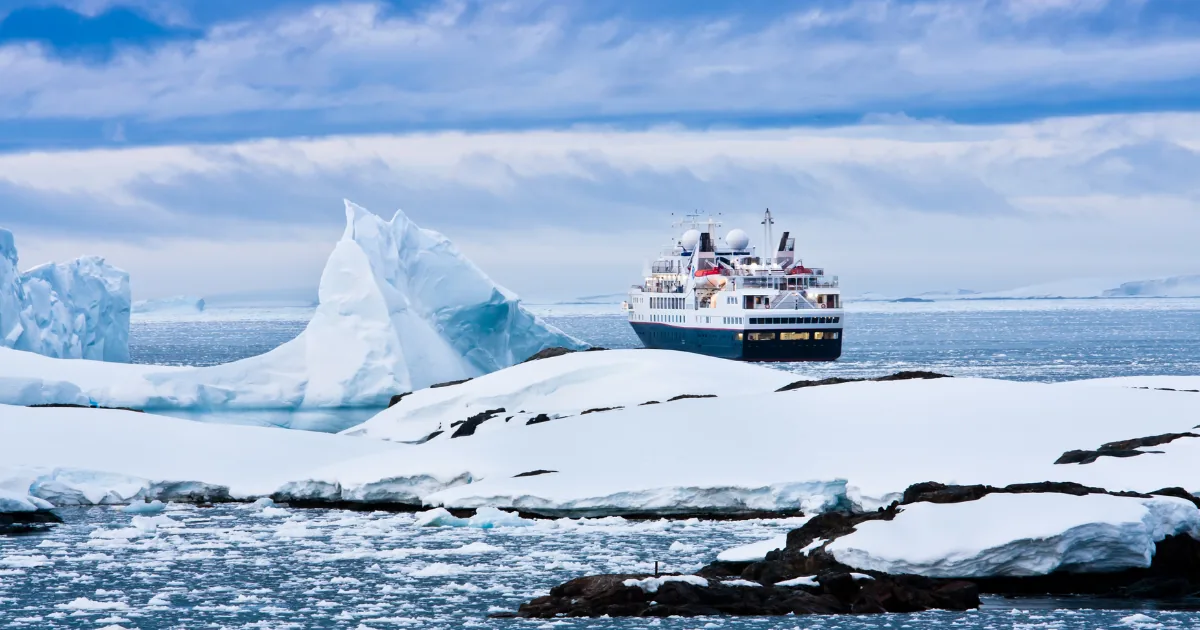 Un barco de expedición navega junto a un iceberg y unas rocas cubiertas de nieve bajo un cielo azul nublado