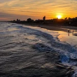 Sunset over a sandy beach with waves rolling in and a town skyline along the shore