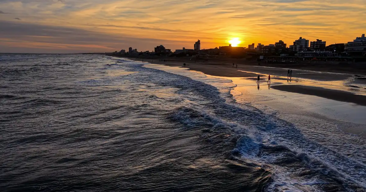 Sunset over a sandy beach with waves rolling in and a town skyline along the shore