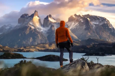 Wanderer in einer orangefarbenen Jacke mit Blick auf die Cuernos del Paine über einem türkisfarbenen See, Torres del Paine, chilenisches Patagonien