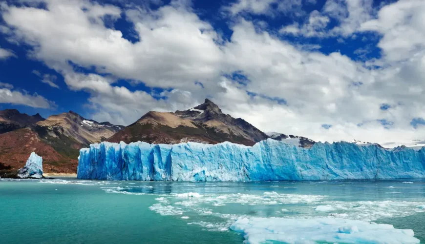 Parete di ghiaccio blu di un ghiacciaio che sfocia in un lago turchese ai piedi di montagne brune, Parco Nazionale Los Glaciares, Santa Cruz, Argentina