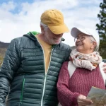 Una pareja de personas mayores sonrientes que sostienen un mapa de senderos mientras dan un paseo por las colinas