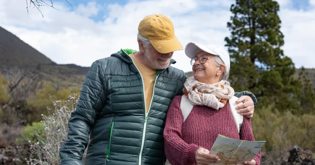 Una pareja de personas mayores sonrientes que sostienen un mapa de senderos mientras dan un paseo por las colinas