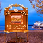 Cartello «Ushuaia, fin del mundo» accanto al porto al tramonto, con le barche per le escursioni ormeggiate dietro, Terra del Fuoco, Argentina