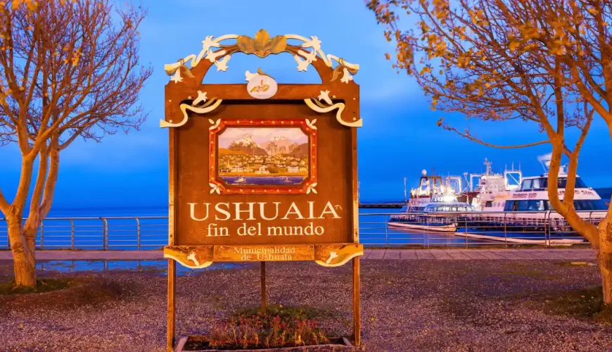 Cartello «Ushuaia, fin del mundo» accanto al porto al tramonto, con le barche per le escursioni ormeggiate dietro, Terra del Fuoco, Argentina