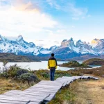 Excursionista en una pasarela de madera frente al macizo del Paine, cubierto de nieve, al otro lado de un lago, en el Parque Nacional Torres del Paine, Chile