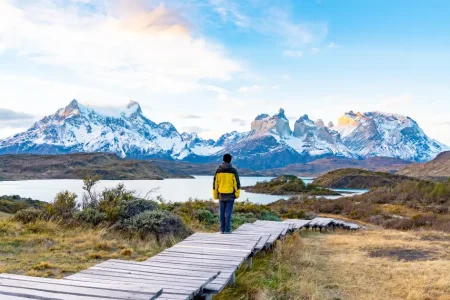 Wanderer auf einem Holzsteg mit Blick auf das schneebedeckte Paine-Massiv jenseits eines Sees, Nationalpark Torres del Paine, Chile