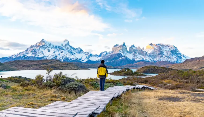 Wanderer auf einem Holzsteg mit Blick auf das schneebedeckte Paine-Massiv jenseits eines Sees, Nationalpark Torres del Paine, Chile