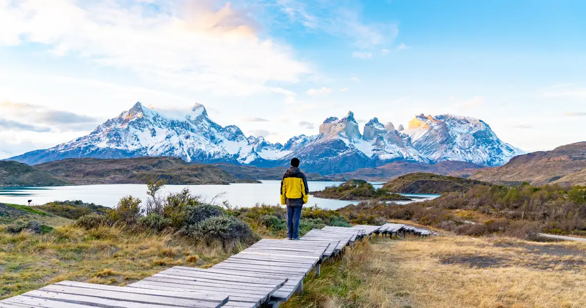Excursionista en una pasarela de madera frente al macizo del Paine, cubierto de nieve, al otro lado de un lago, en el Parque Nacional Torres del Paine, Chile