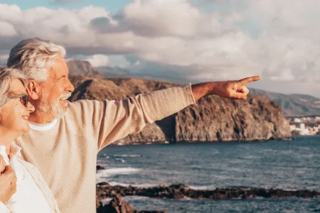 Senior couple smiling and pointing toward a rocky coastline and the sea in warm evening light